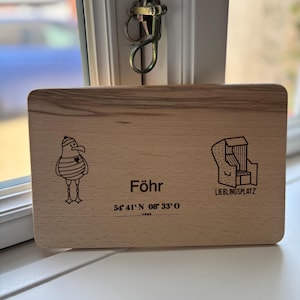 Könnte beinhalten: Rechteckiges Holzschild mit dem Wort "Föhr" und den Koordinaten 54°41'N 08°33'O. Es zeigt eine Cartoon-Möwe und eine Strandkorb-Illustration. Der Text "LIEBLINGSPLATZ" ist ebenfalls vorhanden.