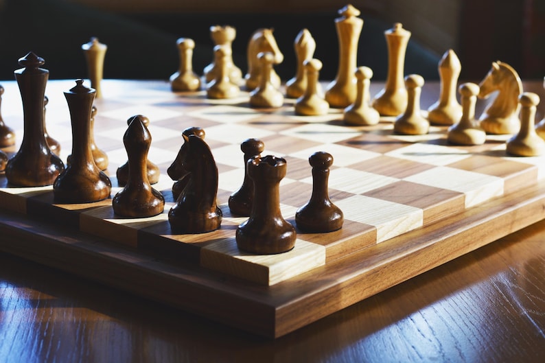 May include: A wooden chessboard with black and white squares. The board is set up with black and white chess pieces. The pieces are arranged in a traditional starting position.