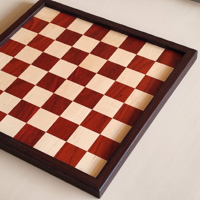 May include: A wooden chessboard with a dark brown frame. The board features a classic checkerboard pattern with alternating red and light-colored squares. The frame is a rich, dark brown, providing a contrasting border to the playing surface.
