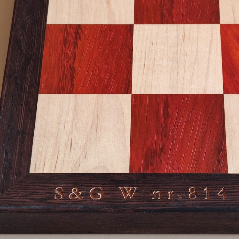 May include: Close-up of a wooden chessboard with alternating red and light wood squares. The board has a dark brown frame with the engraved text "S & G W nr. 814". The chess board is a classic game.