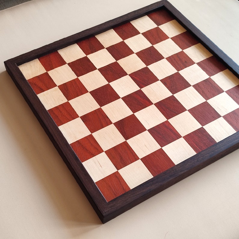 May include: A wooden chess board with a dark brown frame. The playing surface features alternating squares of light wood and reddish-brown wood. The board is square and ready for a game of chess.
