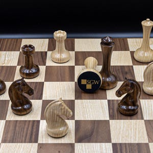 May include: A wooden chessboard with black and white squares. The chess pieces are made of wood and have a dark brown and light brown finish. The pieces are arranged on the board in a game in progress. The board has a logo that says "SGW" in the center.