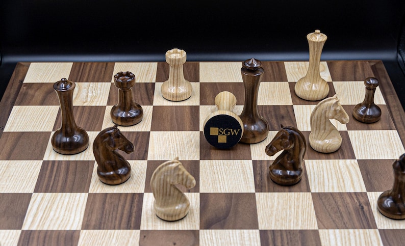 May include: A wooden chessboard with black and white squares. The chess pieces are made of wood and have a dark brown and light brown finish. The pieces are arranged on the board in a game in progress. The board has a logo that says "SGW" in the center.