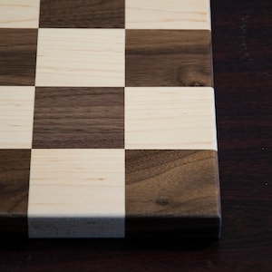 May include: A close-up of a wooden chessboard with alternating squares of light brown maple and dark brown walnut wood. The board is unfinished and has a natural wood grain.