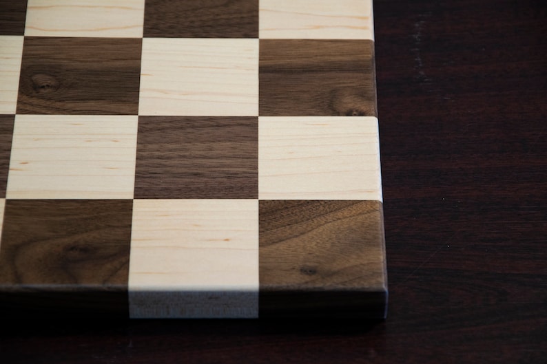 May include: A close-up of a wooden chessboard with alternating squares of light brown maple and dark brown walnut wood. The board is unfinished and has a natural wood grain.