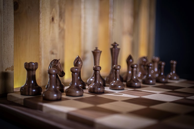 May include: A wooden chessboard with dark brown and light brown squares. The chess pieces are made of dark wood and are arranged in a starting position. The pieces include a king, queen, rooks, bishops, knights, and pawns.