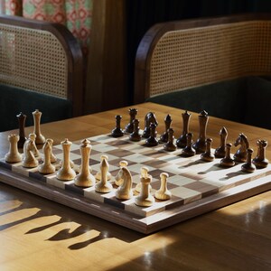 May include: A wooden chessboard with black and white squares. The board is set up with black and white chess pieces. The board is on a wooden table with a natural wood finish.