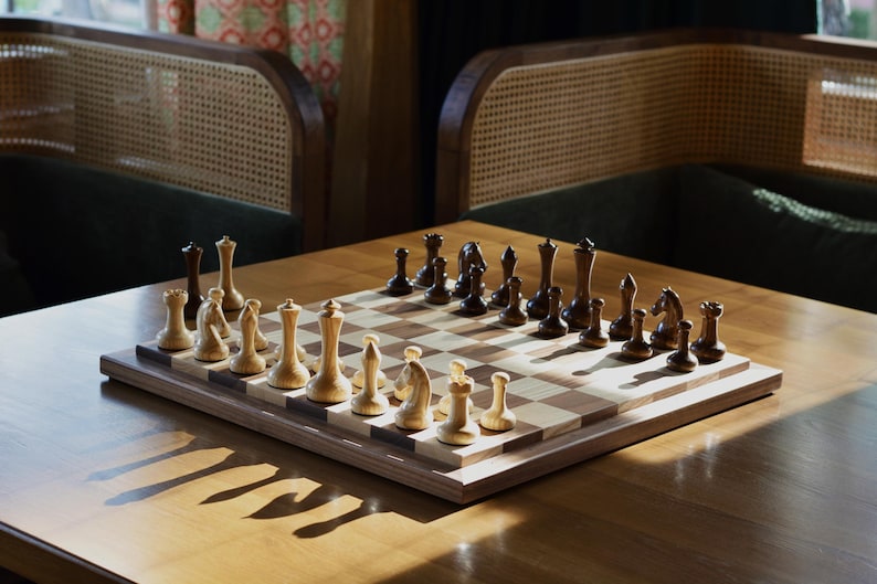 May include: A wooden chessboard with black and white squares. The board is set up with black and white chess pieces. The board is on a wooden table with a natural wood finish.