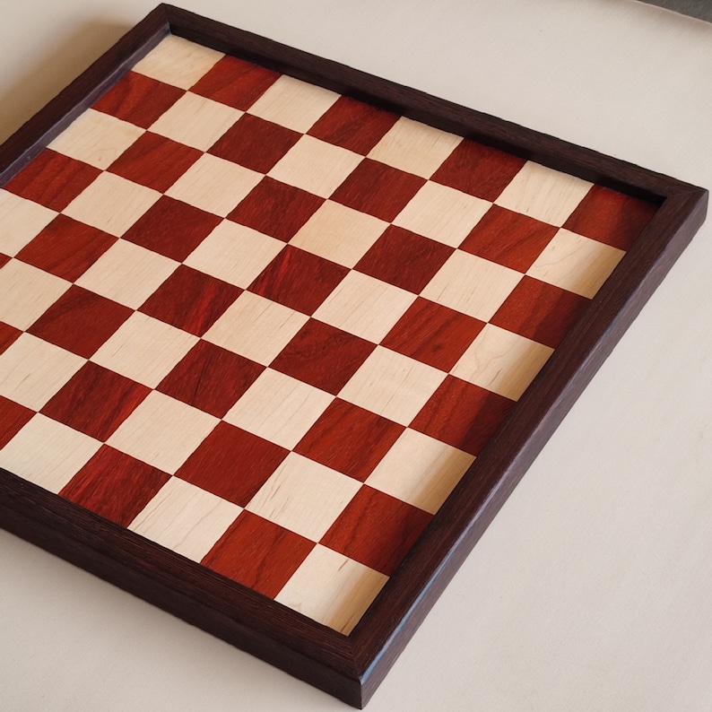 May include: A wooden chess board with a dark brown frame. The playing surface features a classic checkerboard pattern of alternating red and light wood squares. The board is angled, showcasing the detailed craftsmanship.