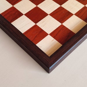 May include: A wooden chessboard with a dark brown frame. The squares alternate between a light wood color and a reddish-brown hue. The board is set against a neutral background.