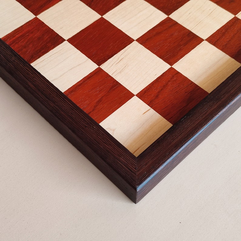 May include: A wooden chessboard with a dark brown frame. The squares alternate between a light wood color and a reddish-brown hue. The board is set against a neutral background.
