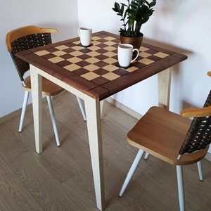 Könnte beinhalten: Ein quadratischer Holztisch mit einer Tischplatte im Schachbrettmuster. Zwei weiße Stühle mit Holzsitzen und -lehnen stehen auf beiden Seiten des Tisches. Eine Topfpflanze und zwei Tassen stehen auf dem Tisch.