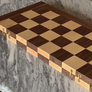 Op de afbeelding: Een opvouwbaar houten schaakbord met een klassiek patroon van afwisselende vierkanten in donkerbruin walnoothout en lichtbeige esdoornhout. Het bord is aan de zijkant voorzien van kleine goudkleurige metalen scharnieren om het dubbel te vouwen.