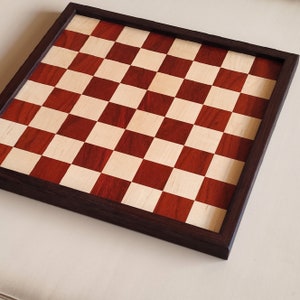 May include: A wooden chessboard with a dark brown frame. The playing surface features alternating red and light-colored squares. The board is square and appears to be made of high-quality wood, suitable for chess games.