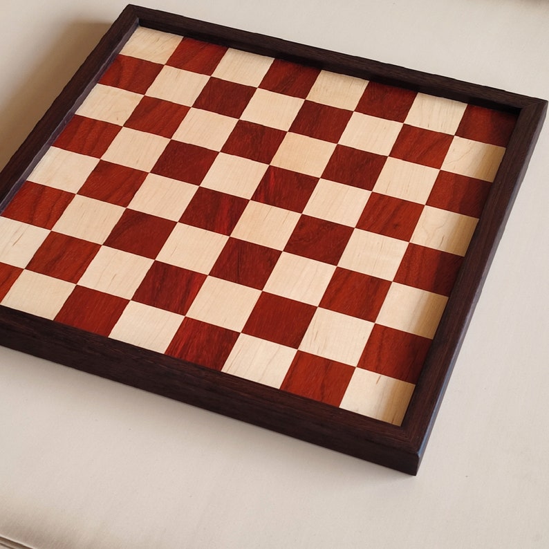 May include: A wooden chessboard with a dark brown frame. The playing surface features alternating red and light-colored squares. The board is square and appears to be made of high-quality wood, suitable for chess games.