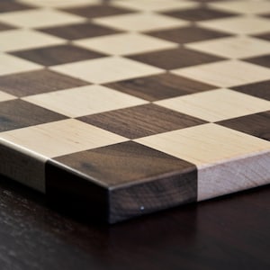 May include: A close-up of a wooden chessboard with alternating squares of light and dark wood. The board is made of maple and walnut wood.