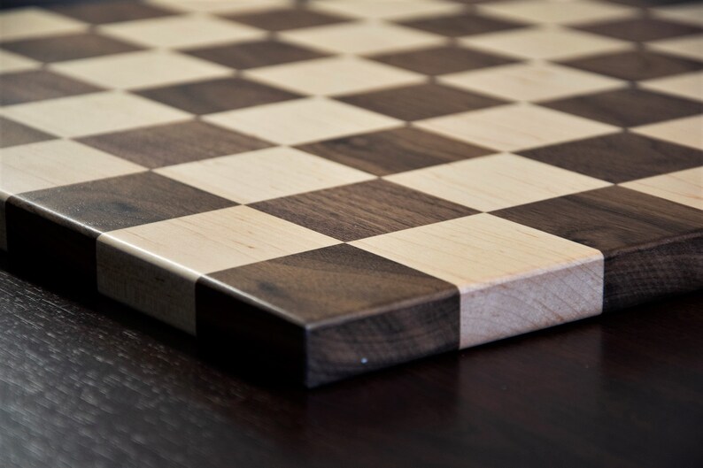 May include: A close-up of a wooden chessboard with alternating squares of light and dark wood. The board is made of maple and walnut wood.