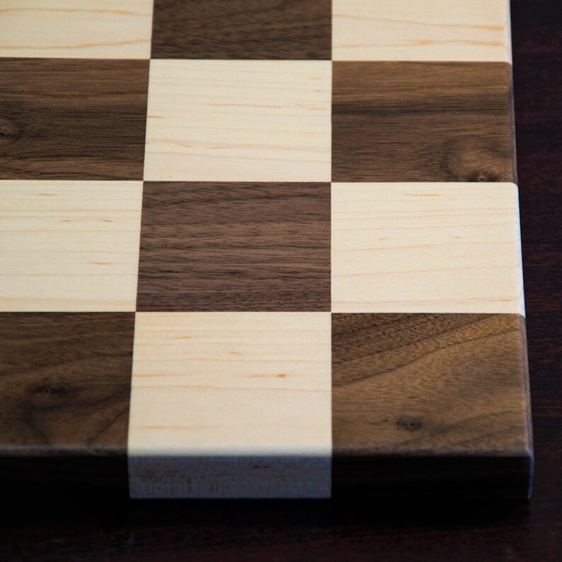 May include: A close-up of a wooden chessboard with alternating light and dark squares. The board is made of maple and walnut wood.