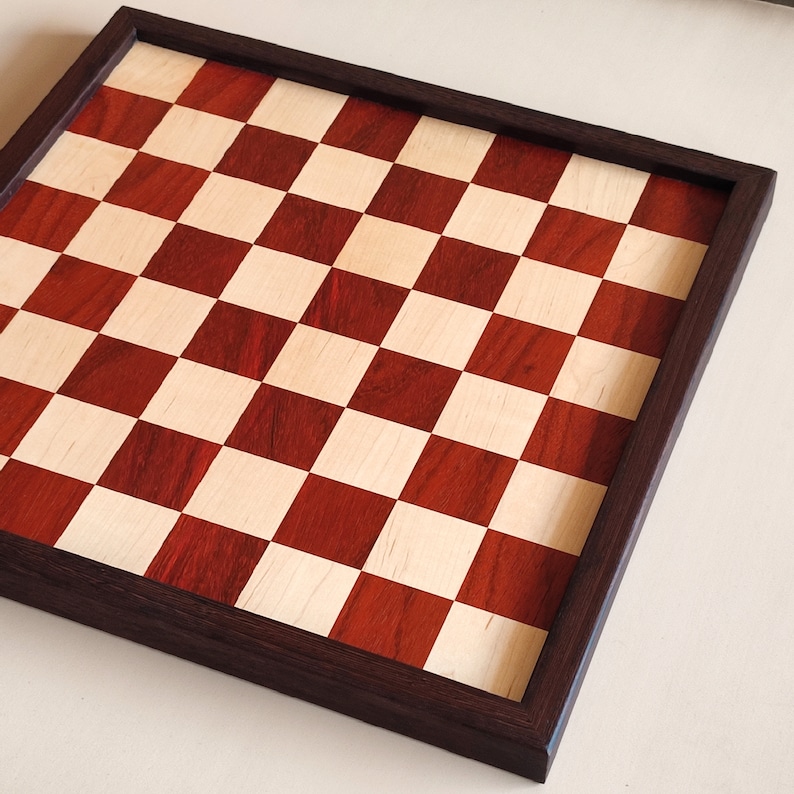 May include: A handcrafted wooden chessboard with a dark brown frame. The playing surface features a classic checkerboard pattern of alternating red and light-colored squares. The board is square and ready for play.
