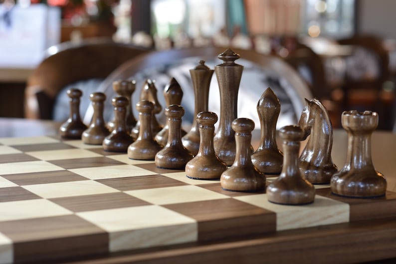 May include: A wooden chessboard with brown and white squares.  The board is set up with a variety of chess pieces, including pawns, rooks, knights, bishops, and a king and queen. The pieces are made of dark wood and are arranged in a traditional chess formation.