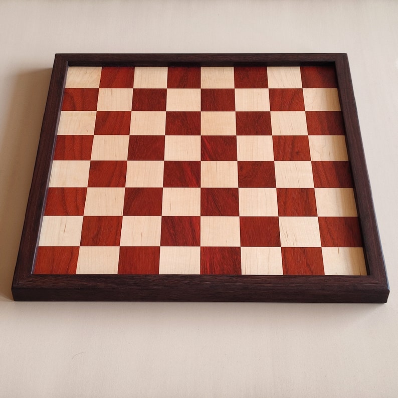 May include: A wooden chessboard with a dark brown frame. The playing surface features alternating squares of light and dark wood, creating a classic checkerboard pattern. The board is designed for strategic gameplay.