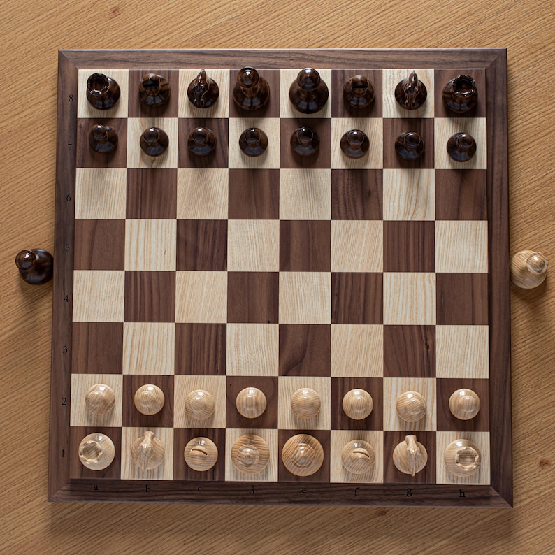 May include: A wooden chessboard with a walnut finish and a light wood inlay. The board is set up for a game of chess with all of the pieces in their starting positions. The pieces are made of wood and have a dark brown finish.