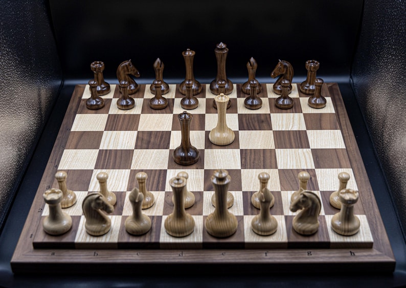 May include: A wooden chessboard with black and white squares. The chess pieces are made of wood and have a dark brown finish. The pieces are arranged in a standard starting position, with the white king and queen in the center of the board. The white king is in check.