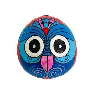 May include: A hand-painted coconut with a blue and red owl design. The owl has large, white eyes with black pupils and a pink beak. The coconut is decorated with swirls and lines in blue and white.