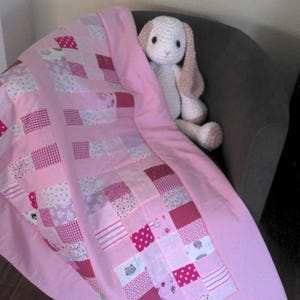 Könnte beinhalten: Eine rosa-weiße Patchwork-Babydecke mit einem gehäkelten weißen Hasen mit rosa Ohren, der darauf sitzt.