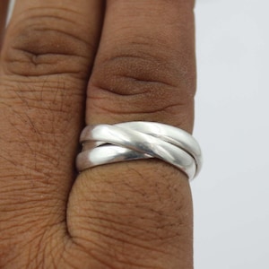 Può includere: Un anello d'argento con un design a tre fasce. Le fasce sono intrecciate, creando un look unico e moderno. L'anello è mostrato su un dito, evidenziando le sue dimensioni e la sua forma. Il metallo lucido riflette la luce, donandogli un aspetto brillante.