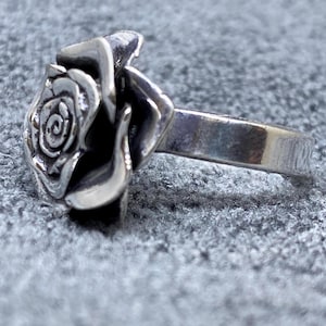 Könnte beinhalten: Ein silberner Ring mit einem detaillierten Rosenmotiv. Die Rose ist der Mittelpunkt, mit aufwendigen Details der Blütenblätter. Das Band ist schlichtes, poliertes Silber. Der Ring ist vor einem strukturierten, grauen Hintergrund platziert.