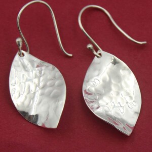 以下が含まれることがあります： ハンマー加工されたテクスチャの、銀色の葉の形をしたイヤリングのペア。 イヤリングはフック留め具で、赤い背景に表示されています。 シンプルでエレガントなデザインです。