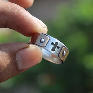 May include: A silver and copper ring with a cross cutout design. The ring has a textured surface and is decorated with small round accents.
