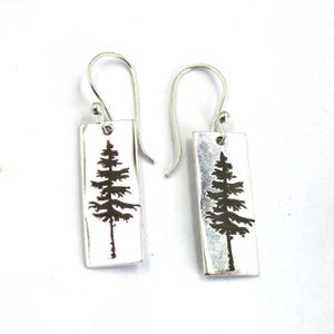 May include: A pair of silver earrings with a rectangular design featuring a black silhouette of a pine tree.