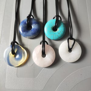 May include: Six gemstone pendant necklaces, each featuring a circular, donut-shaped pendant with a central hole, suspended on a black cord. The pendants display a range of colours: blue, turquoise, pink, white, and yellow, offering a variety of options.