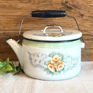 May include: White enamel teapot with a black handle and lid. The teapot features a floral design with orange and blue flowers, and a green band around the middle. The handle is black and the spout is white.