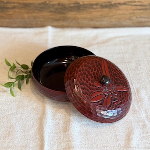 May include: A red lacquered wooden bowl with a matching lid. The lid features a carved floral design and a small, dark knob. The bowl's interior is black, and the exterior has a textured pattern. The bowl is open, revealing its interior.