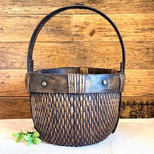 May include: A handcrafted basket with a dark brown woven body and a sturdy black handle. The basket features a decorative metal band around the top, secured with rivets. The basket is designed for carrying items and has a rustic, vintage aesthetic.