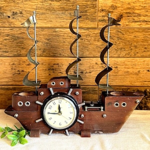 May include: A wooden ship-shaped clock with a clock face in the center and decorative metal sails. The clock has a dark brown finish and is set against a wooden background. The clock's face has black numbers and hands.