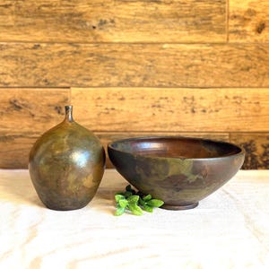 May include: A bronze-colored, pear-shaped vase with a small neck and a matching bowl. The bowl has a wide opening and a rounded base. A small sprig of green leaves sits in front of the bowl. The background is a wooden wall.