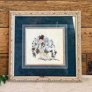 May include: Framed cross-stitch artwork featuring an angel with wings, holding flowers. The angel is dressed in a white gown with floral accents. The artwork is framed in a decorative, ornate, light-colored frame with a dark teal matting.