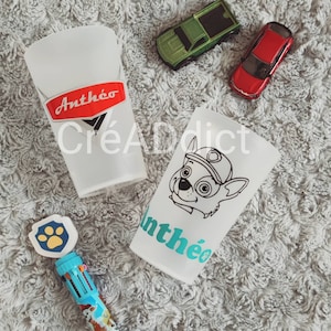 May include: Two white cups with personalized designs. One cup features a red logo with the text "Anth&eacute;o" and the other cup features a black and white cartoon dog with the text "Anth&eacute;o" in teal.