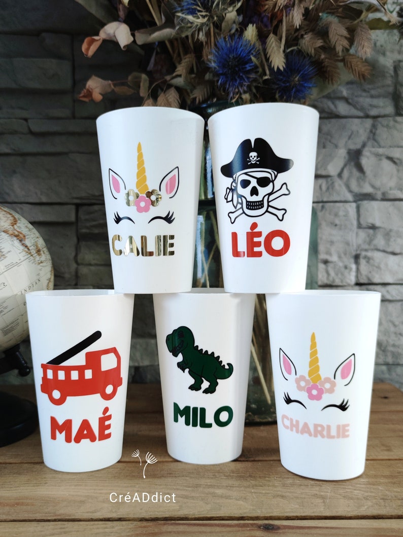 May include: Six white cups with colorful designs and names. The cups feature a unicorn, a pirate skull, a fire truck, a dinosaur, and two more unicorns. The names on the cups are Callie, L&eacute;o, Ma&eacute;, Milo, and Charlie.