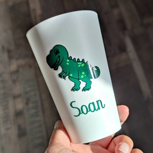 May include: White plastic cup with a green dinosaur graphic and the name "Soan" printed on the side.