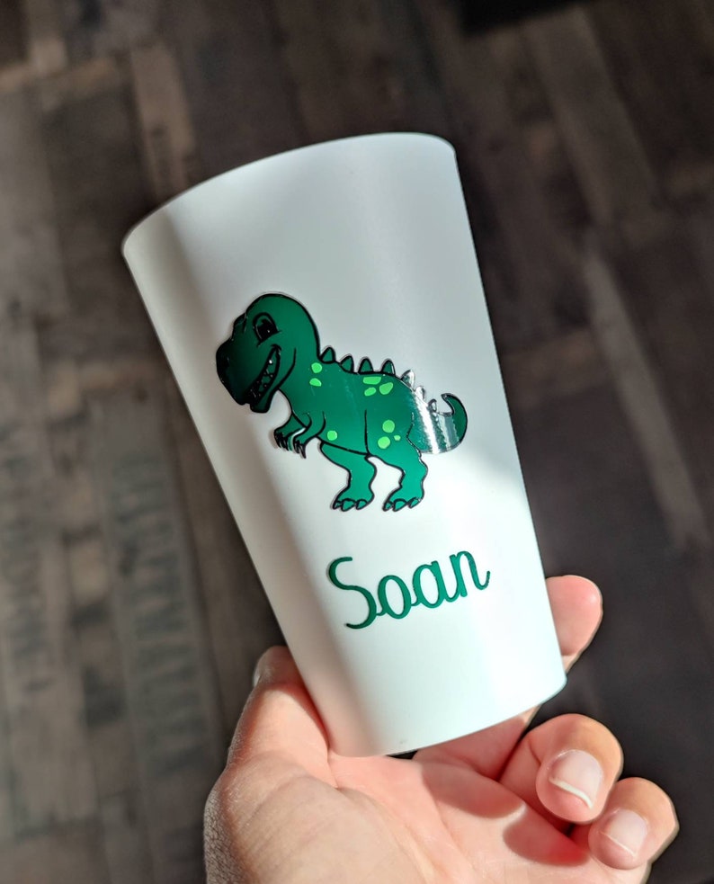 May include: White plastic cup with a green dinosaur graphic and the name "Soan" printed on the side.