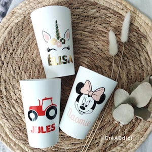 May include: Three white plastic cups with personalized designs. The first cup features a unicorn with the name "ELISA" in gold lettering. The second cup has a red tractor with the name "JULES" in black lettering. The third cup has a pink and black Minnie Mouse design with the name "NAOMIE" in pink lettering.