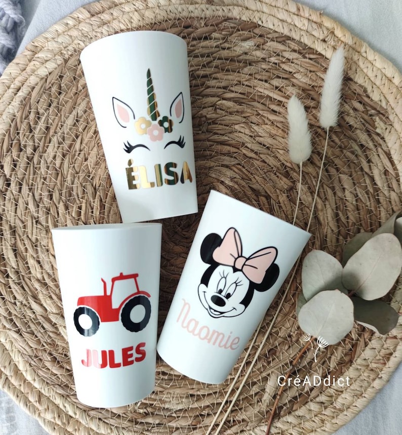 May include: Three white plastic cups with personalized designs. The first cup features a unicorn with the name "ELISA" in gold lettering. The second cup has a red tractor with the name "JULES" in black lettering. The third cup has a pink and black Minnie Mouse design with the name "NAOMIE" in pink lettering.