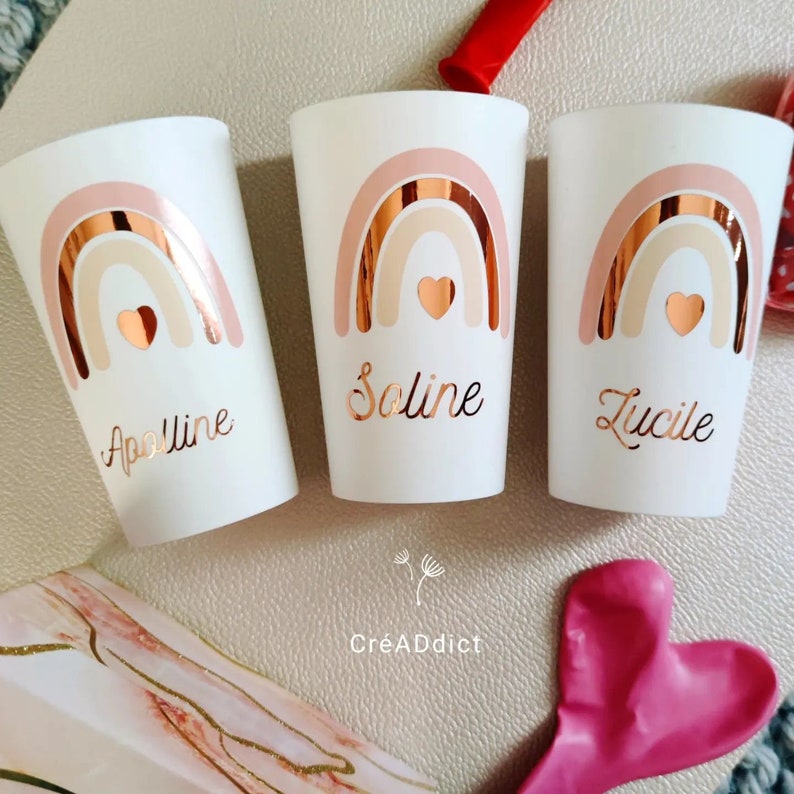 May include: Three white cups with pink and gold rainbow designs and personalized names: Apolline, Soline, and Lucille. The cups are on a white surface with a pink heart balloon and a white box with gold accents.