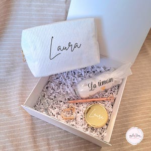 Può includere: Una scatola regalo bianca che contiene una pochette bianca con cerniera con il nome "Laura" stampato su di essa. La scatola contiene anche una piccola bottiglia di vetro con il testo "La témoin" su di essa, un ciondolo a forma di cuore dorato, uno specchio compatto dorato con il nome "Laura" inciso su di esso e una penna rosa oro.