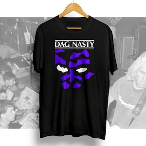 Peut inclure: T-shirt noir avec un motif violet et blanc d'un visage et le texte "DAG NASTY" en blanc.